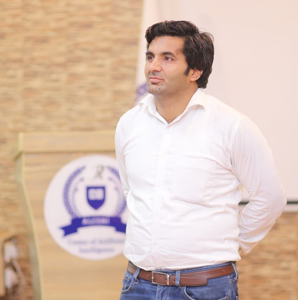 Man in a white shirt and jeans stands in front of a podium with a shield logo.